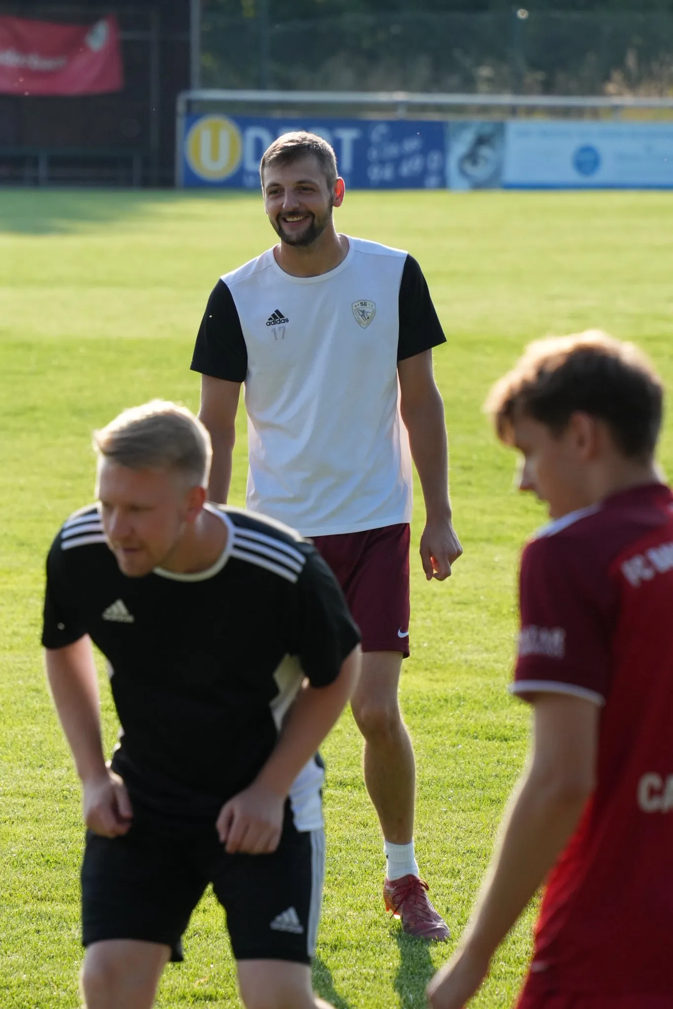 Eine Gruppe von Fußballspielern in Trainingskleidung auf einem Sportplatz, wobei zwei Spieler im Vordergrund zusammen trainieren und einer im Hintergrund lächelt. Der Rasen ist gut gepflegt und es gibt Werbebanner im Hintergrund.