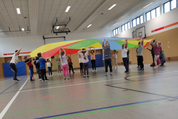 Eine Gruppe von Kindern und Erwachsenen steht in einer Turnhalle mit einem bunten, großen Parasol. Die Teilnehmer halten den Parasol hoch, während sie an einer spielerischen Aktivität teilnehmen, umgeben von Basketballkörben und Turnmatten.