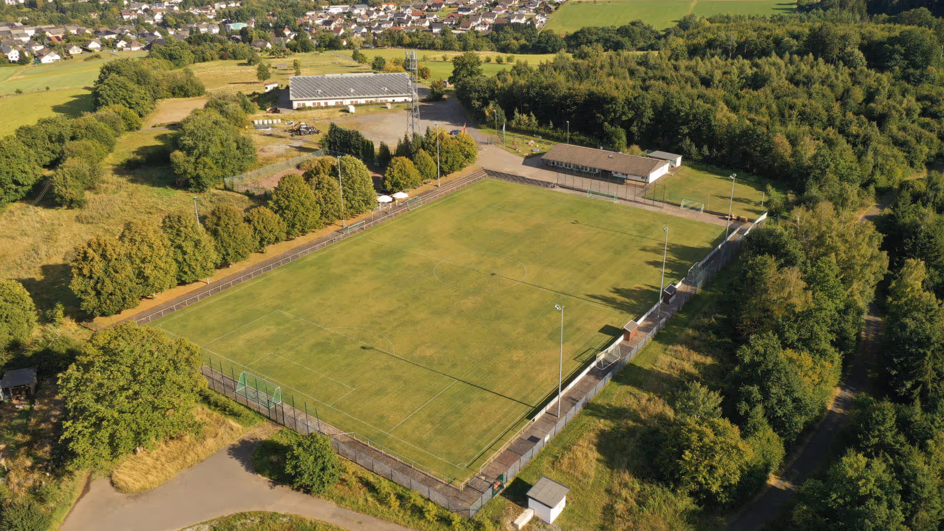 Luftaufnahme eines großen, gut gepflegten Fußballplatzes umgeben von Bäumen und einem angrenzenden Gebäude. Im Hintergrund sind Wohngebäude und Felder sichtbar.